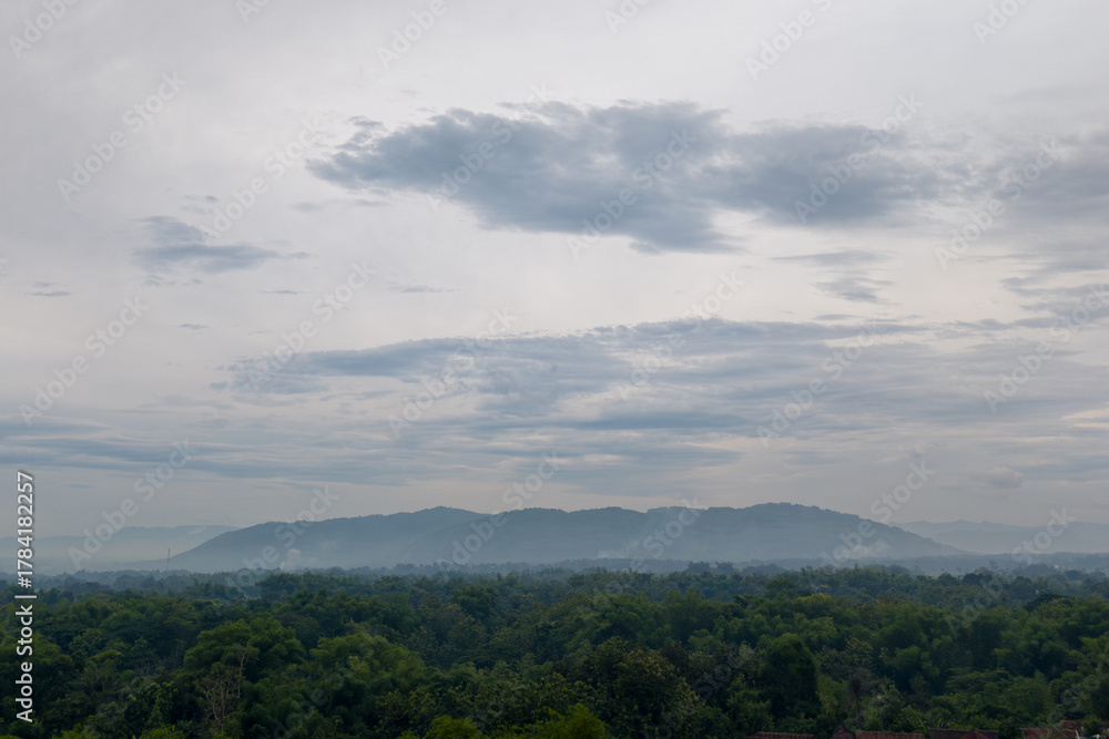 Obraz premium Panoramic landscape of a hazy, majestic volcano mountain dominating the horizon under a dramatic, cloudy morning or evening sky, overlooking a dense rural forest.