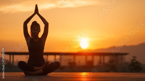 A silhouette of a woman in a black tank top and leggings, meditating on a wooden deck with a mountainous background and a vibrant sunset sky.