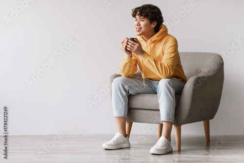 Fotografie Cheerful Asian man sits comfortably in a gray armchair, sipping coffee