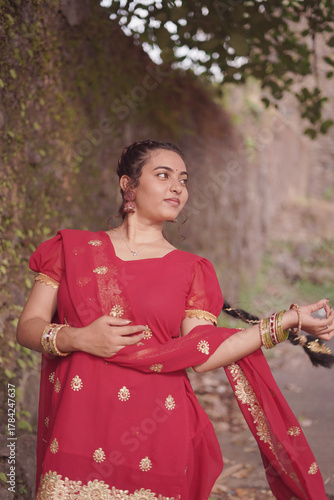 A young woman wearing a traditional red salwar kameez with golden embroidery stands outdoors near an old stone wall covered with natural moss