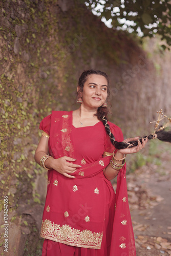A young woman wearing a traditional red salwar kameez with golden embroidery stands outdoors near an old stone wall covered with natural moss