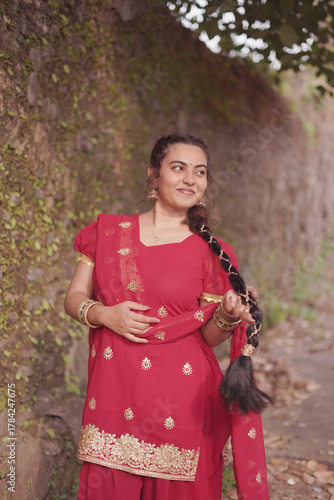 A young woman wearing a traditional red salwar kameez with golden embroidery stands outdoors near an old stone wall covered with natural moss