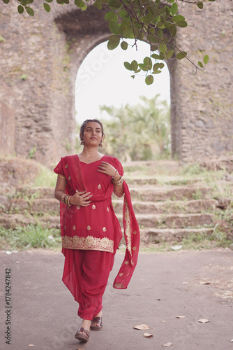A young woman wearing a traditional red salwar kameez with golden embroidery stands outdoors near an old stone wall covered with natural moss