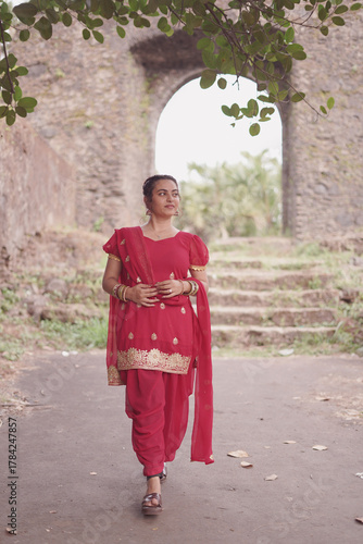 A young woman wearing a traditional red salwar kameez with golden embroidery stands outdoors near an old stone wall covered with natural moss