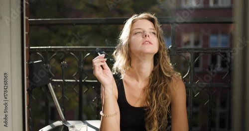 Girl relaxing on balcony enjoying cigarette while basking in the sun outside an apartment in the city during a pleasant afternoon