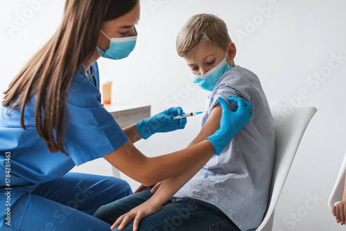 A preteen boy is getting vaccinated against the covid-19 virus in a modern clinic. He is sitting and wearing a mask while a nurse administers the vaccine in his arm.