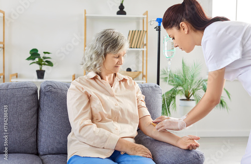 Young friendly nurse or caregiver help sick senior elderly woman patient in receiving IV drip infusion and vitamin injection therapy sitting on sofa in medical clinic. Health care concept.