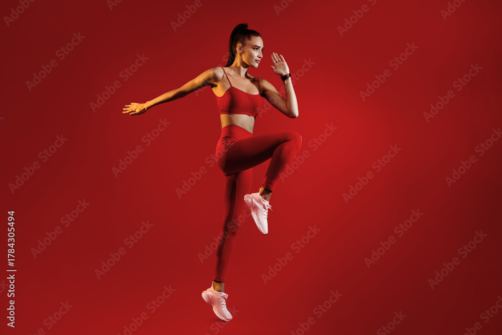 Obraz premium A fitness enthusiast showcases athleticism with an energetic pose in striking red workout attire. The backdrop enhances the intensity of her movement, highlighting strength and grace.