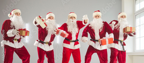 Dancing funny cute group wearing red Santa Claus suits humorous New Year fun with gift box, performance holiday party, energetic dance, playful and lively studio fun surprising joy spirit team
