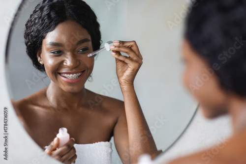 Photography Beautiful young african american lady smiles at her reflection while applying facial serum under her eyes, enjoying the smoothness of her skin as part of her daily beauty routine