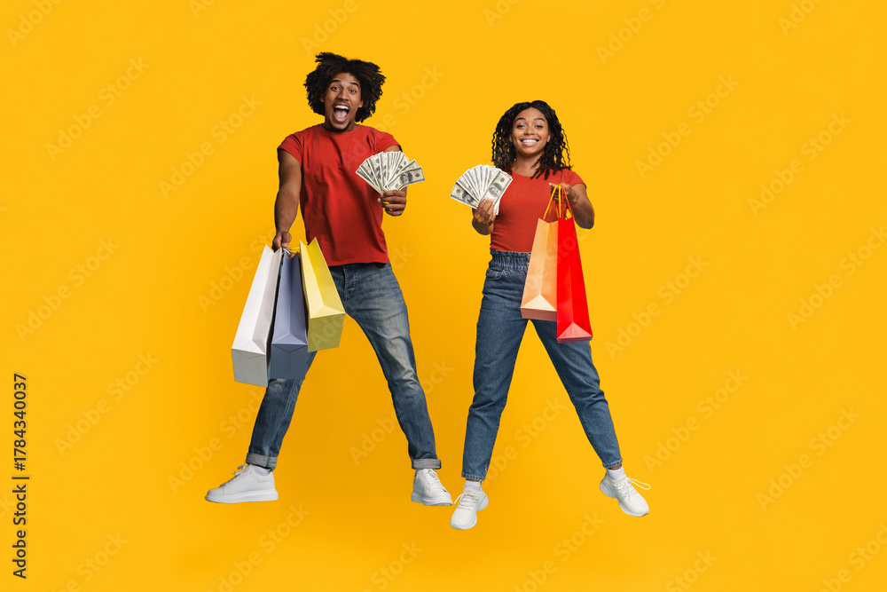 Fototapeta premium Two joyful friends jump against a bright yellow background, holding cash and colorful shopping bags. Their enthusiasm shows they are having a fun shopping day together.