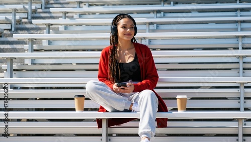 Young woman enjoys music with headphones and phone while sitting on bleachers with coffee cups