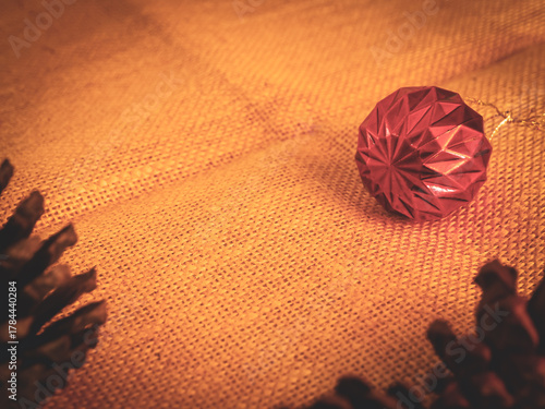 Red Christmas ball with dried pine cones on brown sackcloth background in warm vintage style. Cozy and rustic festive decoration perfect for holiday cards, winter season design, or minimalist