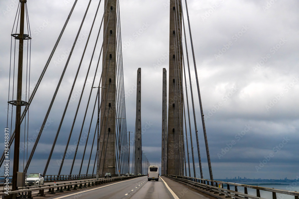 Obraz premium Vehicles driving along the Oresund bridge, a prominent cable stayed structure connecting Denmark and Sweden under a cloudy sky. 