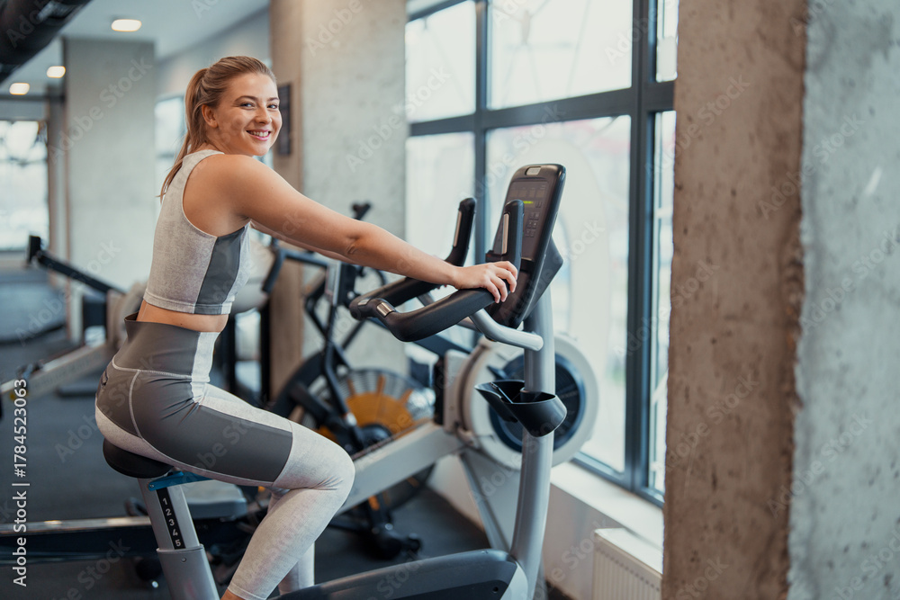 Fototapeta premium Smiling young Caucasian woman exercising on stationary bike in modern gym with natural light