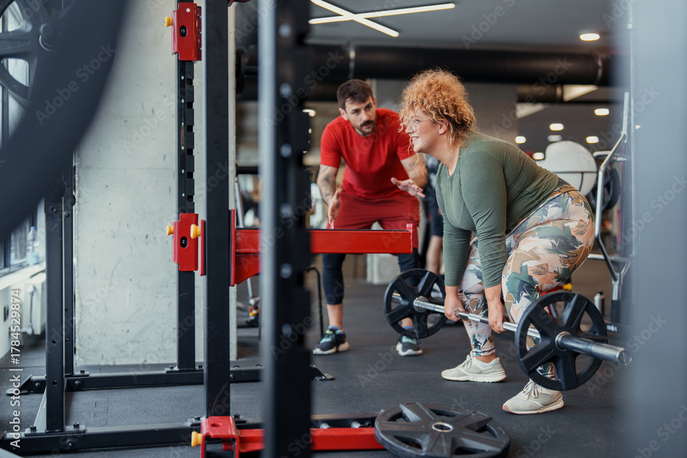 Fototapeta premium Two People Exercising Indoors Woman Deadlifting with Man Coaching in Gym