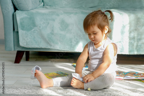 A one-year-old girl sitting on the floor among children's books trying to put on her socks