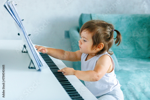 A one-year-old girl at the piano looks with interest at the sheet music on the music stand
