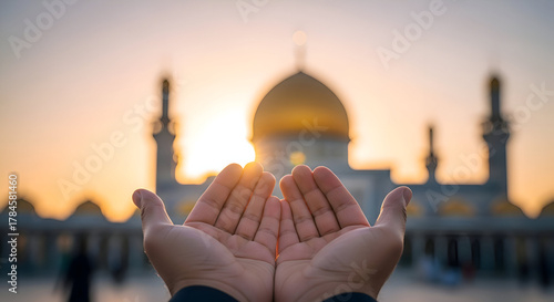 A person's hands held open in a gesture of prayer towards a grand mosque with a golden dome at sunrise.