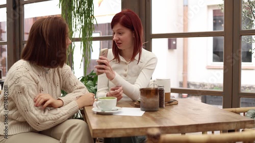 Two coworkers sit together one proudly shows something on her phone with a satisfied smile while the other looks on with envy trying to hide her feelings but affected by the moment