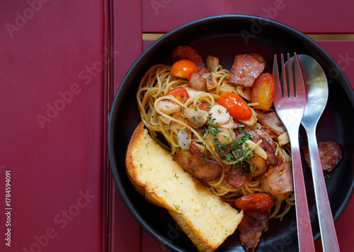 Top view food of spaghetti bacon with fried garlic and tomato serve on black dish with toast on red wooden table.
