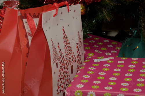 Close-up of Christmas gifts under a tree with red bags and pink floral wrapping paper. Festive holiday mood and seasonal decoration concept.