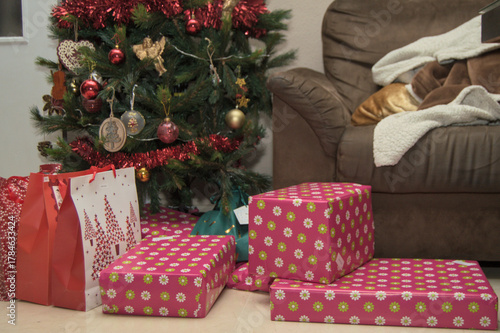 Cozy living room with red-decorated Christmas tree and gifts beside a brown sofa. Warm festive home interior for Christmas and winter holidays.