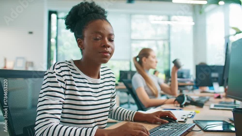 Beautiful African American woman in striped shirt smiling and looking directly at camera while sitting at desk. Female employee distracted from tasks on table in bright shared office workspace.