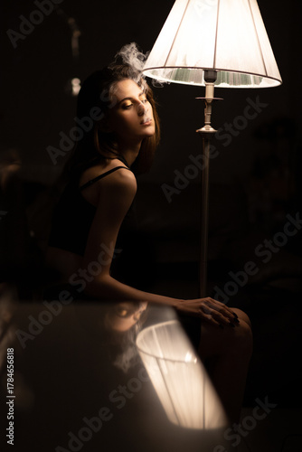 Female portrait. Young woman is smoking in dark room near the lamp. Low key photography.