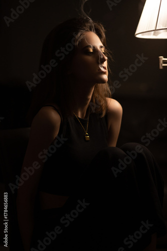 Female portrait. Young woman is smoking in dark room near the lamp. Low key photography.