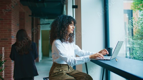 Curly-haired Caucasian woman sitting near window working on laptop focused on screen. Wearing white shirt and brown trousers. Modern corridor with brick walls and plants. Blurred colleague passing by.