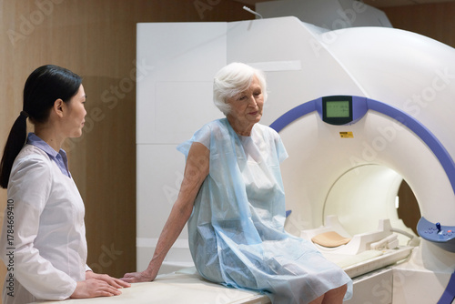 Senior Caucasian woman sitting on bed of MRI machine after being scanned, young Asian female nurse assisting her