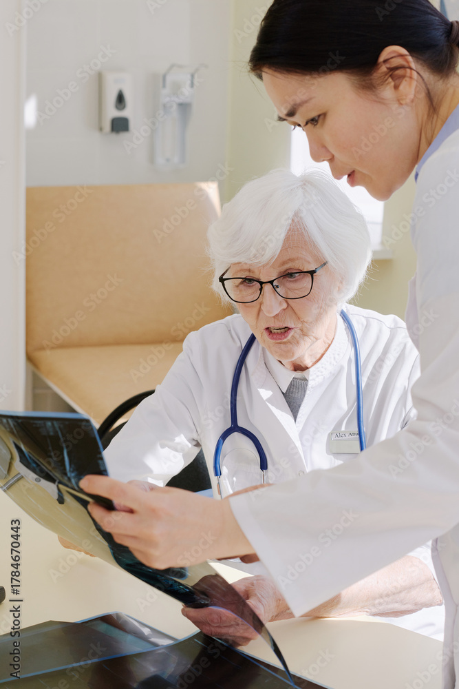 Obraz premium Mature female physician analyzing conditions of patient by looking at his X-ray after MRI scan and consulting with younger assistant