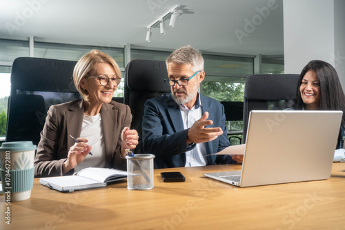 Three busy happy middle aged professional business man and two women executive leaders team using laptop working on computer at work desk having conversation on financial project at meeting in office.