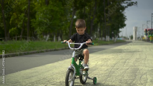 Three year old boy smiles and looks at the camera while riding a green kids bicycle on asphalt in the park with a forest background