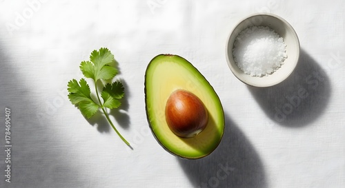 Fresh avocado half with seed, cilantro leaves, and a small bowl of salt on a white surface, highlighting healthy eating and natural ingredients