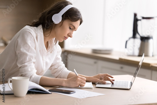 Young woman student wearing headphones writing watching webinar on notebook