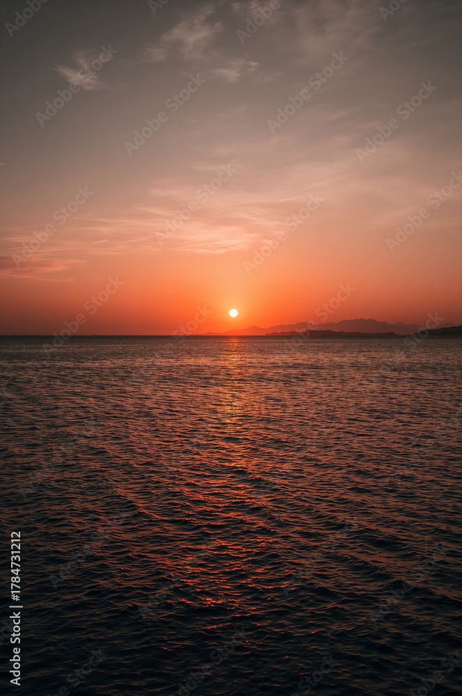 Naklejka premium Stunning sunset paints the ocean with fiery orange hues reflecting on calm water, creating a serene and majestic coastal panorama.