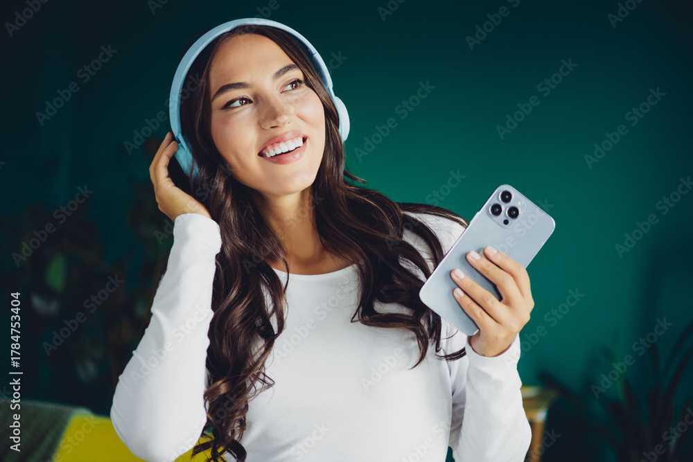 Naklejka premium Happy young woman with headphones at home smiling while using phone in a modern living room setting for relaxed everyday moments