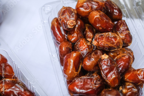 Close-up of fresh dates piled in a clear plastic container on a white surface with warm lighting.