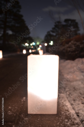 A glowing white paper lantern sits on a snowy sidewalk at night with blurred lights and trees in the background.