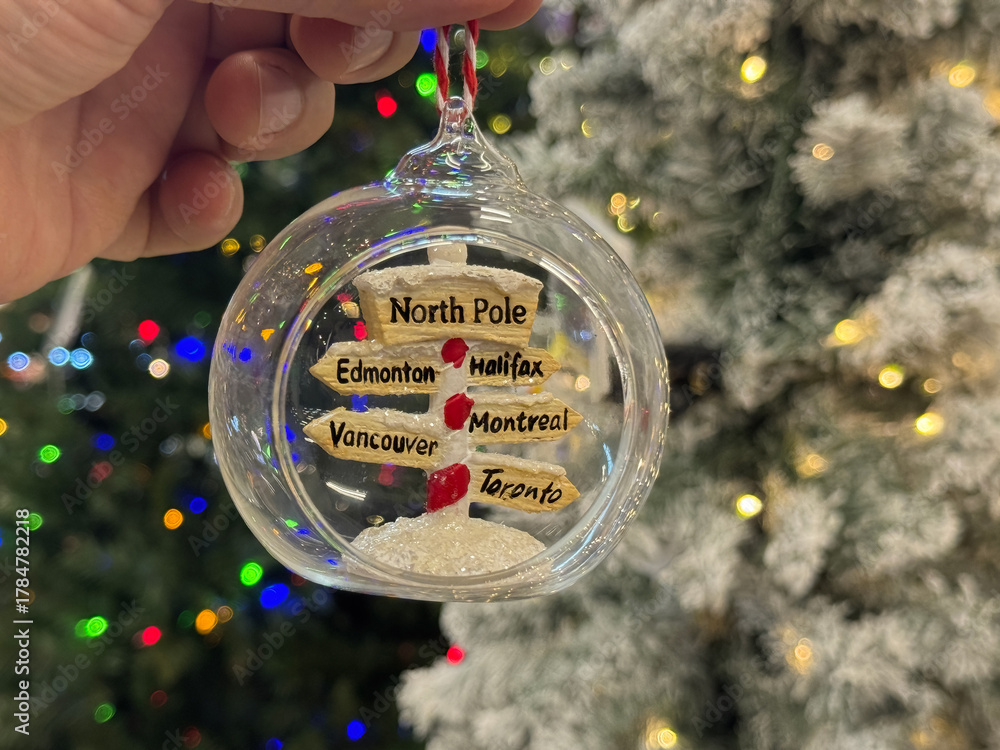 Fototapeta premium Hand-held glass Christmas ornament with a miniature North Pole signpost and wooden city direction signs over a snowy base.