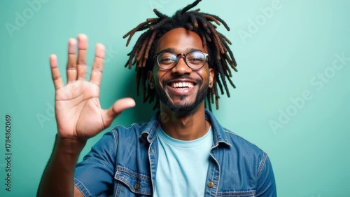 African American man with glasses and dreadlocks smiles while waving hand, showcasing friendly demeanor, camera performs a subtle zoom in to capture joyful expression