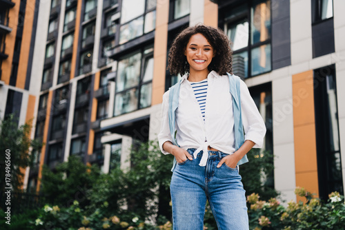 Young woman student stands in front of modern city apartments smiling confidently in casual urban attire conveying lifestyle and travel vibe for stock photography