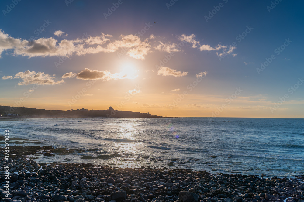 Fototapeta premium Summer sunset in Shimen District, New Taipei City, Taiwan, with golden sunlight reflecting on calm sea, rocky shore, and distant hill silhouette.