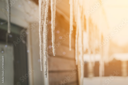 Sharp icicles hanging down from the roof of the residential building against sunny background. Transparent icicles on facade of building, dangerous icicles melt on rooftop, risk of injury
