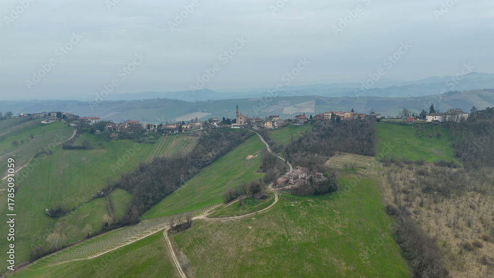 Obraz premium Panoramic vista of the rolling Piacenza hills in Italy revealing clusters of rural architecture and verdant landscapes under a hazy sky with distant mountain ranges and a small village nestled on the