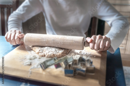 Hands-on festive baking. A person rolling out brown cookie dough with a wooden pin on a board with holiday cutters. Traditional family cooking activity.