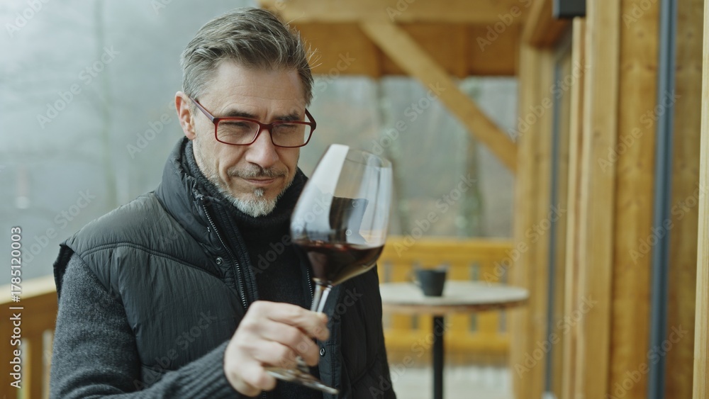 Obraz premium Senior winemaker enjoying aroma of red wine in glass, standing at wooden balcony of winery. Mature man drinking, tasting and analyzing quality of wine.