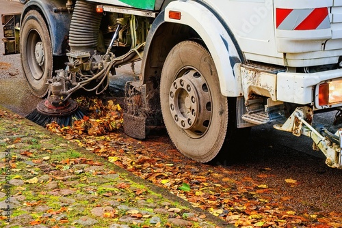 Street sweeper cleaning fallen autumn leaves on a wet cobblestone road. Municipal cleaning vehicle working during fall season.